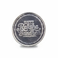 A round silver-colored metallic coin or medallion engraved with 'HAPPY Raksha Bandhan 2021' in stylized lettering, placed on a textured black background.