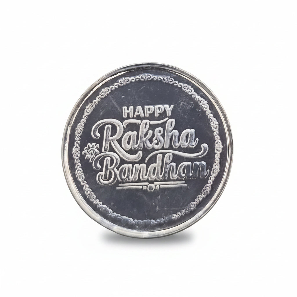 A round silver-colored metallic coin or medallion engraved with 'HAPPY Raksha Bandhan 2021' in stylized lettering, placed on a textured black background.