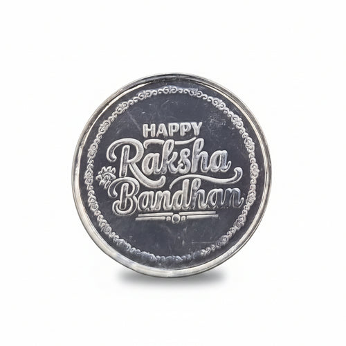 A round silver-colored metallic coin or medallion engraved with 'HAPPY Raksha Bandhan 2021' in stylized lettering, placed on a textured black background.