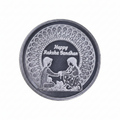 A round silver-toned metal token or button featuring an engraved design with two children holding hands, a floral pattern border, and the text 'Happy Rakha Bandhan' on a black center background.